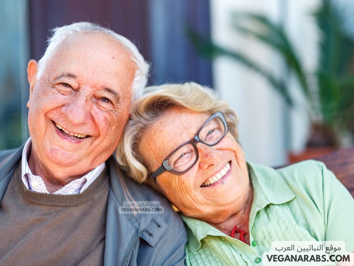 Elderly man and woman smiling warmly, seated closely with heads touching. The background is blurred with hints of greenery, conveying a joyful and relaxed mood.